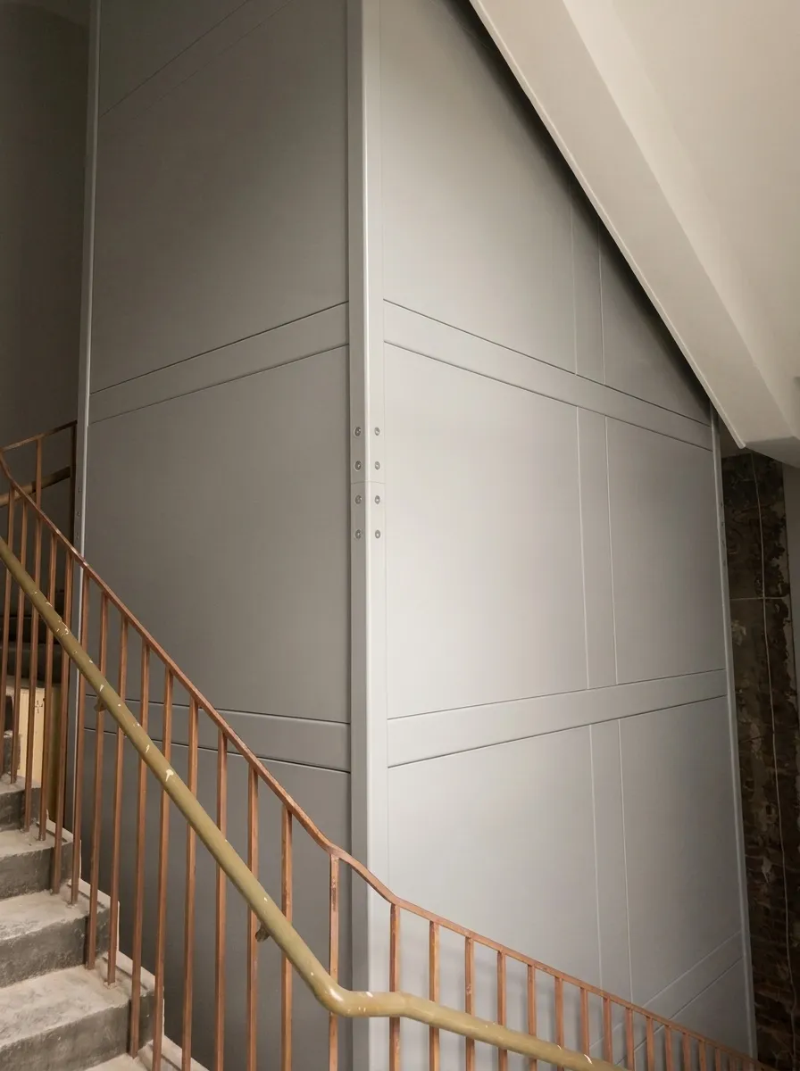 Szyb windy obudowany szarymi panelami stalowymi w klatce schodowej z metalową balustradą
