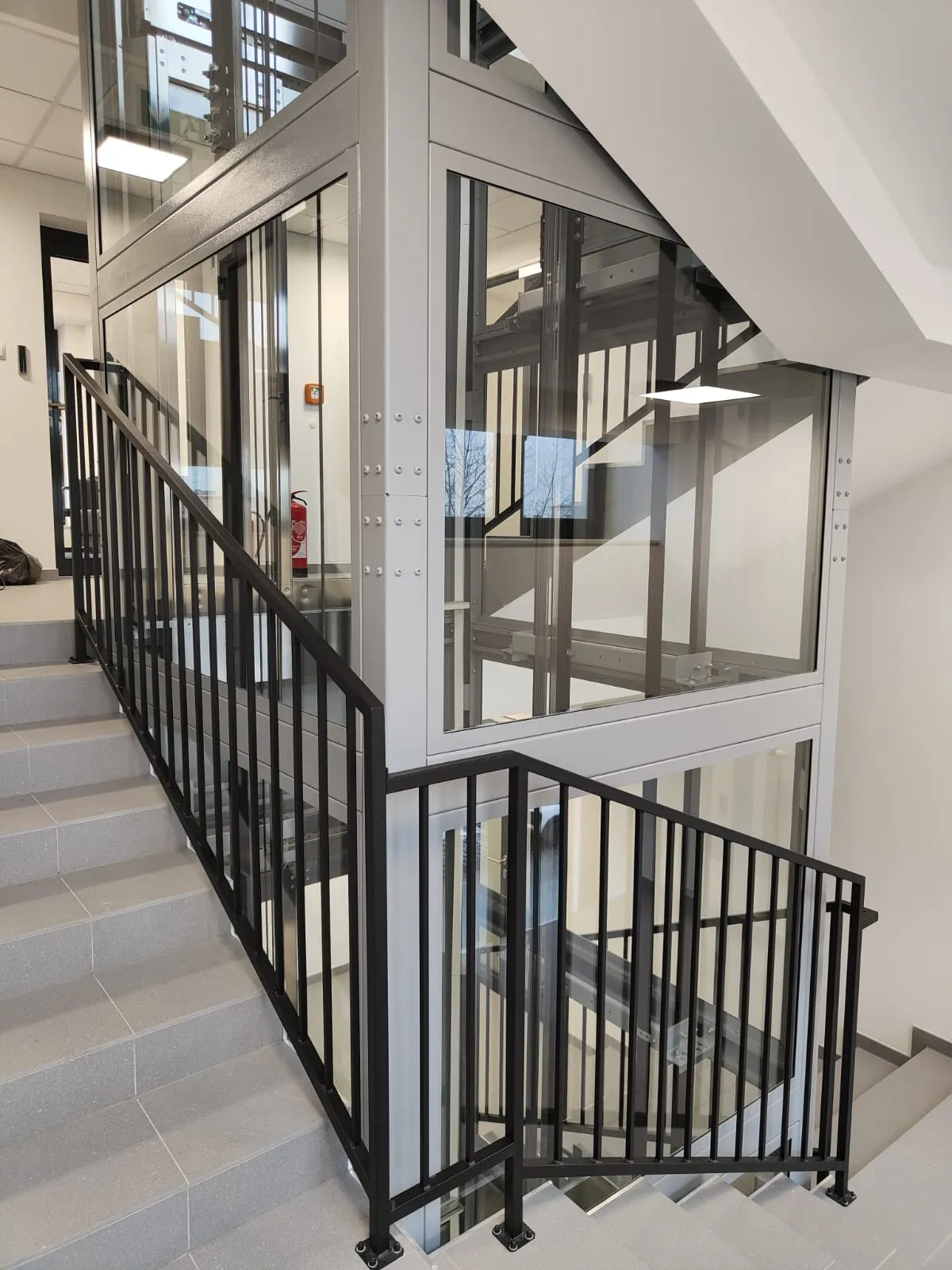 Jasnoszary przeszklony szyb windy w klatce schodowej z czarnymi balustradami — realizacja w budynku publicznym