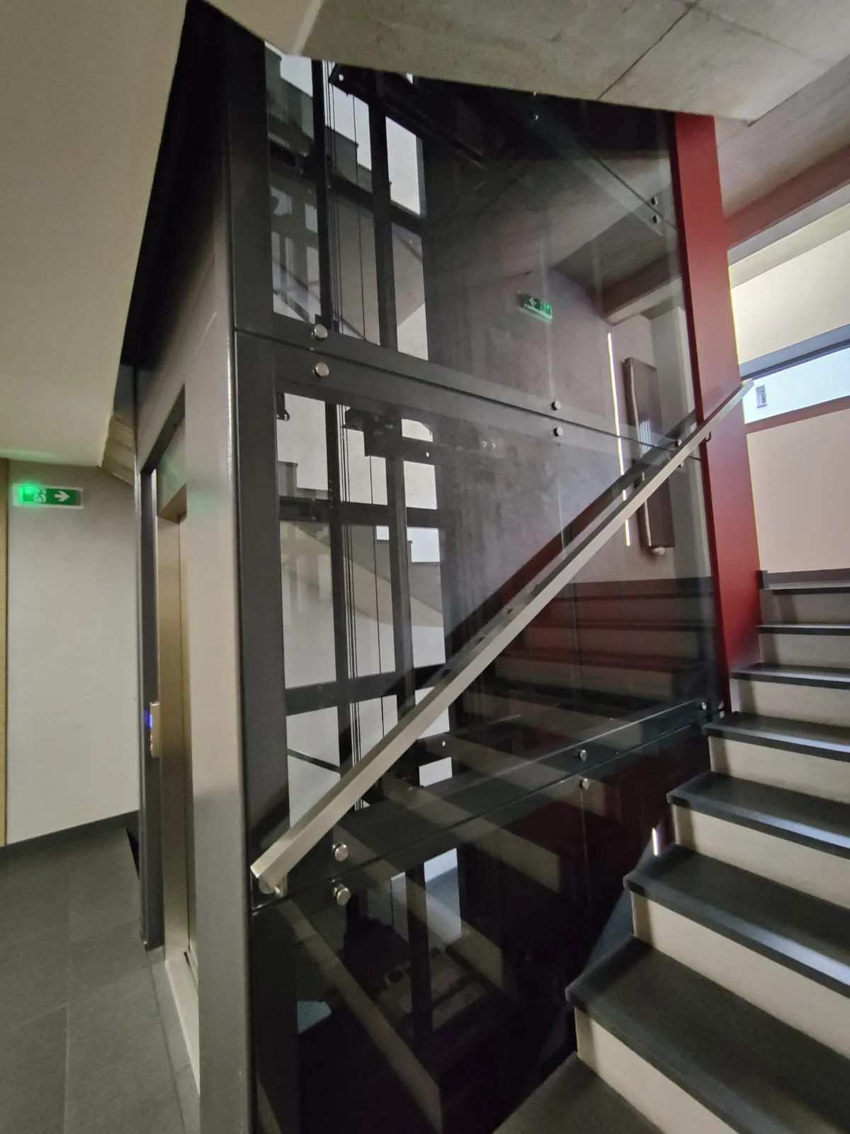 Przeszklony szyb z poręczą biegnącą wzdłuż schodów, za szkłem liny i prowadnice
